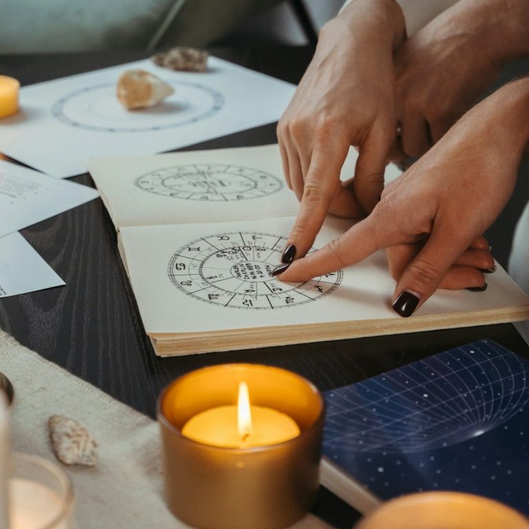 Astrolojik Semboller ve Notlar Açık bir not defteri üzerindeki notlara ve sembollere işaret eden eller, mumlarla çevrili